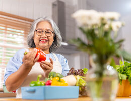 A woman in a kitchen looks at a red bell pepper.