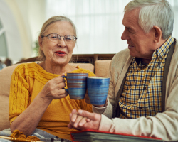  An older couple clinks their mugs together while holding hands on a couch.