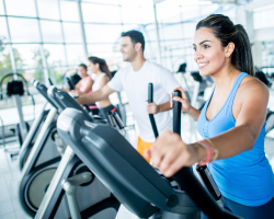 A woman on an elliptical machine with other gym patrons in the background.