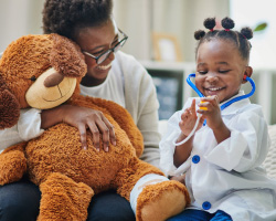 A woman holding a large teddy bear laughs with a young girl in a doctor costume