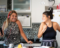Two smiling women in a kitchen. One wears headphones and holds a phone. The other dances.