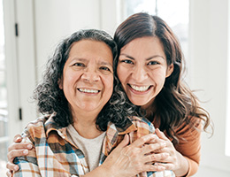 An adult woman standing close behind her mom with her hands on her mom's arms. 