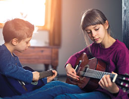 A young girl and boy sit on the floor playing guitars.