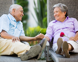 An older woman holding a rose and an older man clasp hands while sitting on lawn furniture.