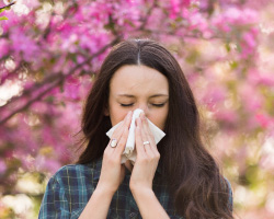 A woman stands in front of a cherry tree while holding a tissue to her nose.