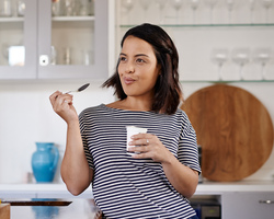 A woman stands in a kitchen eating some yogurt.