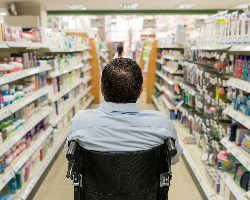 A man using a wheelchair, seen from behind in the toiletries aisle of a grocery store.
