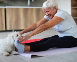 A seated woman stretches toward her toes as a small white dog pokes his head between her feet.