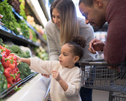 A young girl takes a red pepper from a grocery display as her parents help.